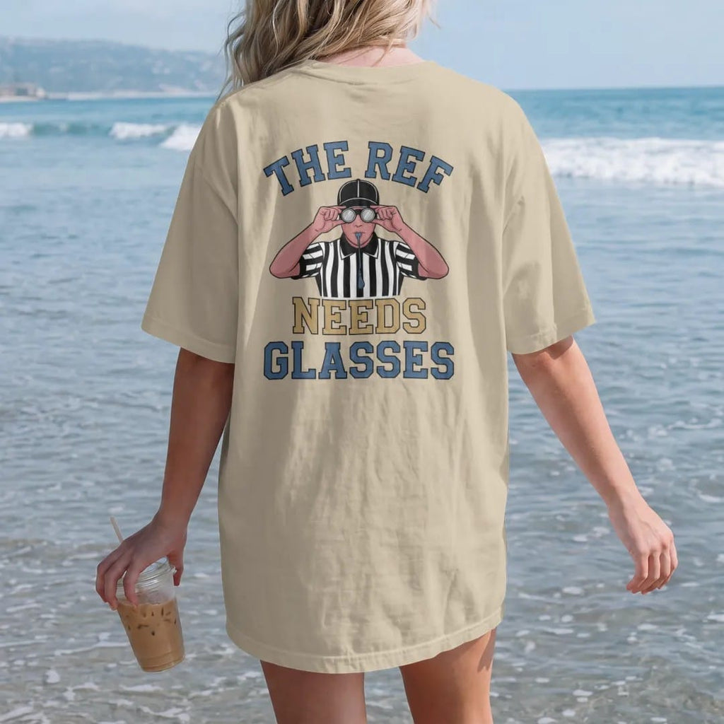 Woman on the beach in a Sandstone tee, back view: a referee in stripes squinting through oversized glasses with a whistle in his mouth, with "THE REF" and "NEEDS GLASSES" in chunky rounded 70s varsity letters, printed in faded stadium green, washed…