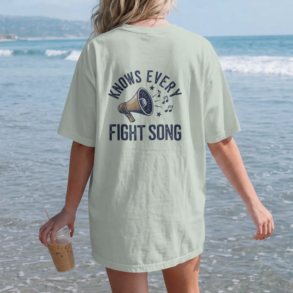 Woman on the beach in a Bay tee, back view: a megaphone with musical notes and stars bursting out, with "KNOWS EVERY" and "FIGHT SONG" in chunky 1970s swash script, printed in faded stadium green, washed brick red, faded navy and mustard with a…