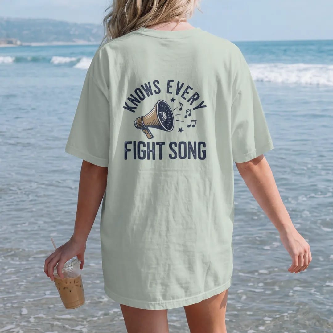Woman on the beach in a Bay tee, back view: a megaphone with musical notes and stars bursting out, with "KNOWS EVERY" and "FIGHT SONG" in chunky 1970s swash script, printed in faded stadium green, washed brick red, faded navy and mustard with a…