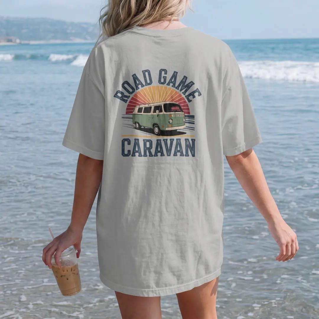 Woman on the beach in a Granite tee, back view: a vintage van covered in pennants driving down a highway, with "ROAD GAME" and "CARAVAN" in chunky 70s slab-serif letters, printed in faded stadium green, washed brick red, faded navy and mustard with…