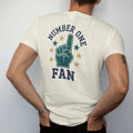 Man wearing a Ivory tee, seen from the back: a giant foam finger surrounded by stars, with "NUMBER ONE" and "FAN" in chunky rounded 70s varsity letters, printed in faded stadium green, washed brick red, faded navy and mustard with a cracked, worn…