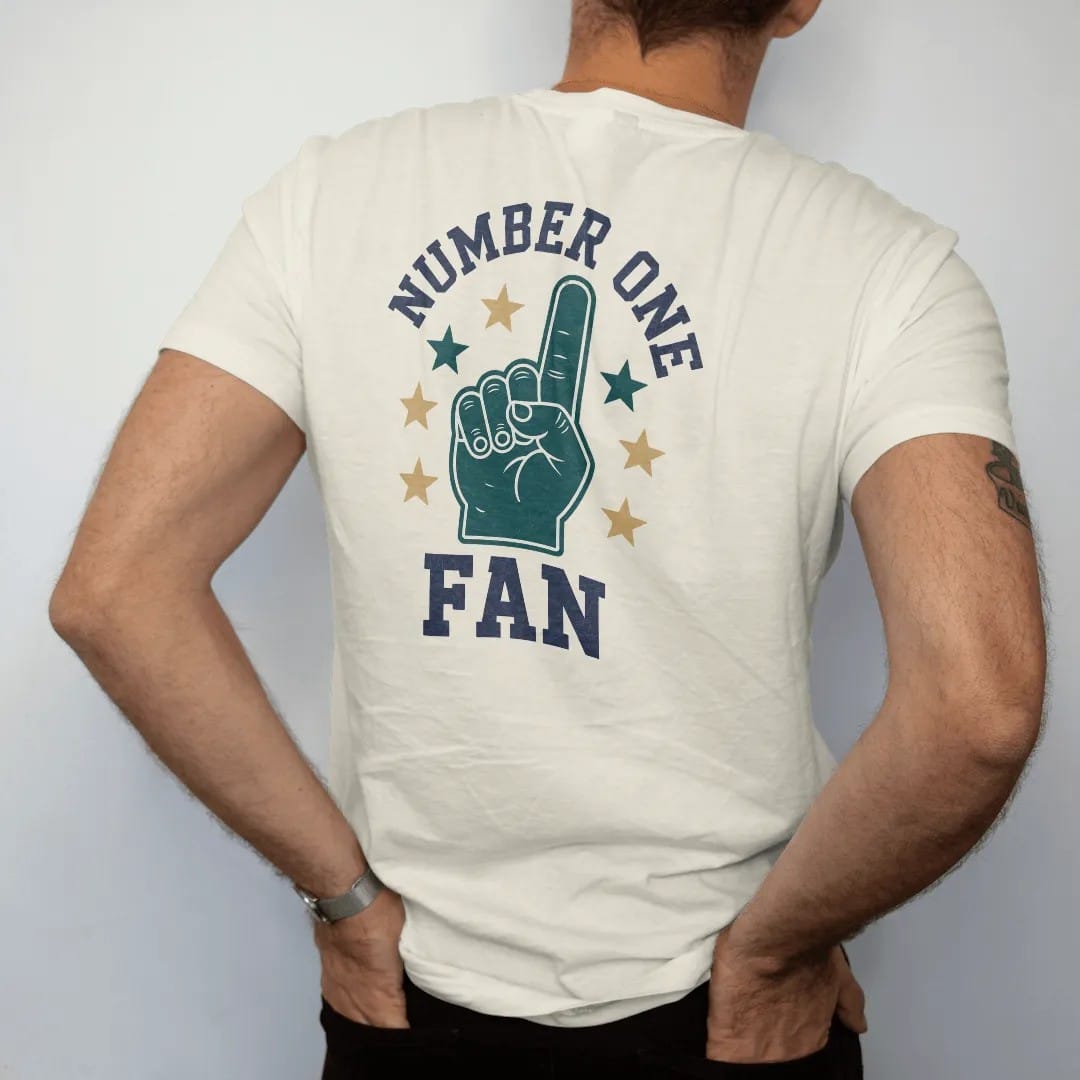 Man wearing a Ivory tee, seen from the back: a giant foam finger surrounded by stars, with "NUMBER ONE" and "FAN" in chunky rounded 70s varsity letters, printed in faded stadium green, washed brick red, faded navy and mustard with a cracked, worn…
