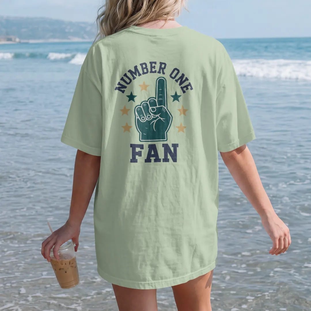 Woman on the beach in a Light Green tee, back view: a giant foam finger surrounded by stars, with "NUMBER ONE" and "FAN" in chunky rounded 70s varsity letters, printed in faded stadium green, washed brick red, faded navy and mustard with a cracked,…