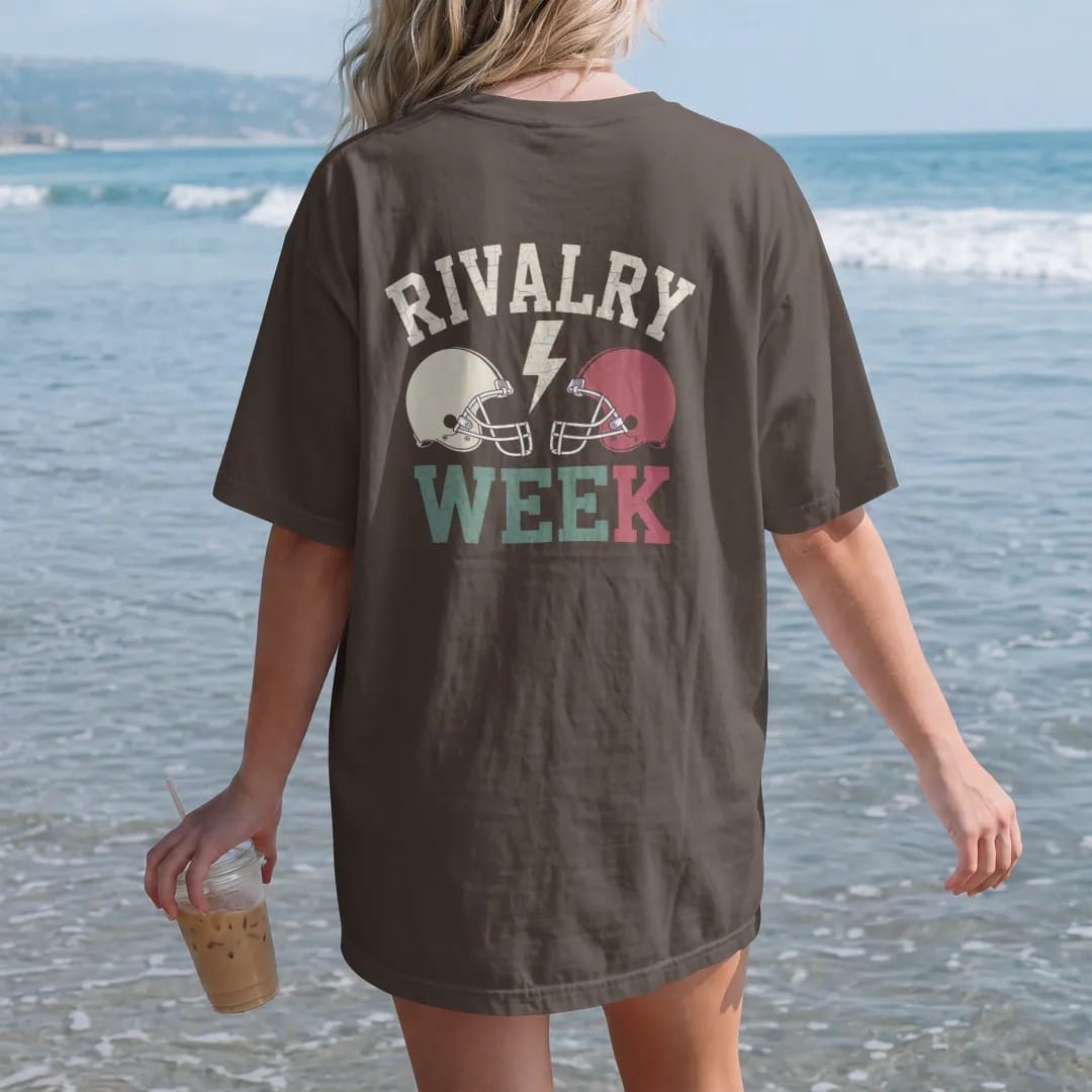 Woman on the beach in a Espresso tee, back view: two plain football helmets facing off with a lightning bolt between them, with "RIVALRY WEEK" in chunky rounded 70s varsity letters, printed in faded bleacher cream, mustard, washed tomato red and…