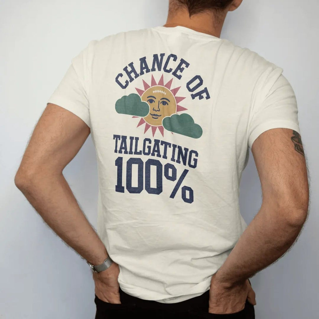 Man wearing a Ivory tee, seen from the back: a retro TV weather icon of a sun with a football face and puffy clouds, with "CHANCE OF" and "TAILGATING" and "100%" in chunky 70s slab-serif letters, printed in faded stadium green, washed brick red,…