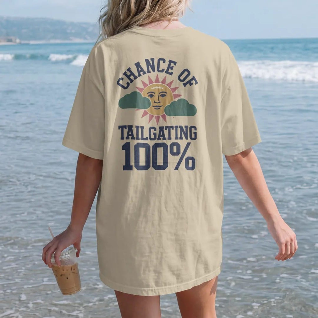 Woman on the beach in a Sandstone tee, back view: a retro TV weather icon of a sun with a football face and puffy clouds, with "CHANCE OF" and "TAILGATING" and "100%" in chunky 70s slab-serif letters, printed in faded stadium green, washed brick…