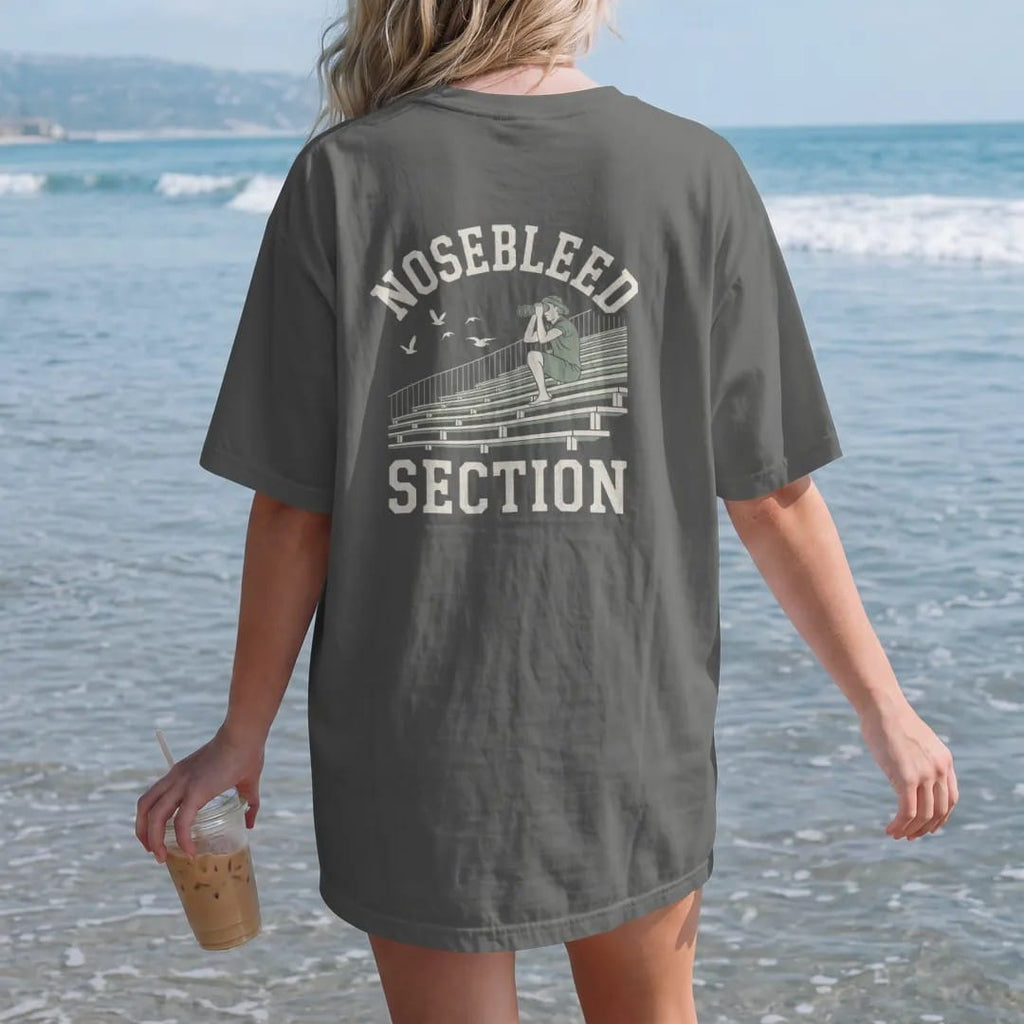 Woman on the beach in a Pepper tee, back view: steep bleacher seats high in a stadium with a fan using binoculars and birds flying past, with "NOSEBLEED" and "SECTION" in chunky rounded 70s varsity letters, printed in faded bleacher cream, mustard,…