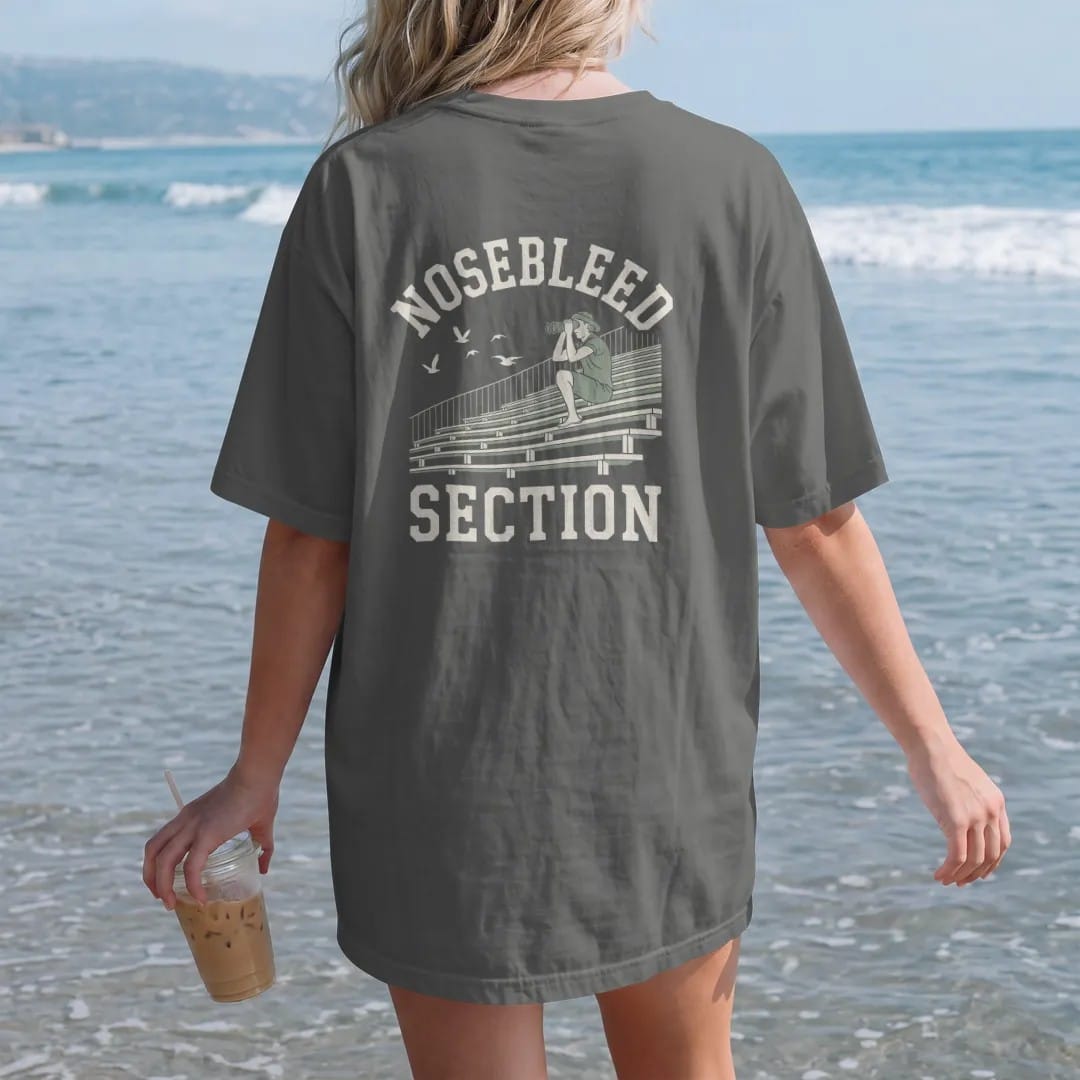 Woman on the beach in a Pepper tee, back view: steep bleacher seats high in a stadium with a fan using binoculars and birds flying past, with "NOSEBLEED" and "SECTION" in chunky rounded 70s varsity letters, printed in faded bleacher cream, mustard,…
