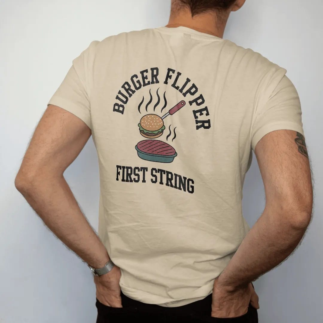 Man wearing a Sandstone tee, seen from the back: a spatula flipping a burger patty into the air with grill smoke, with "BURGER FLIPPER" and "FIRST STRING" in chunky rounded 70s varsity letters, printed in faded stadium green, washed brick red,…