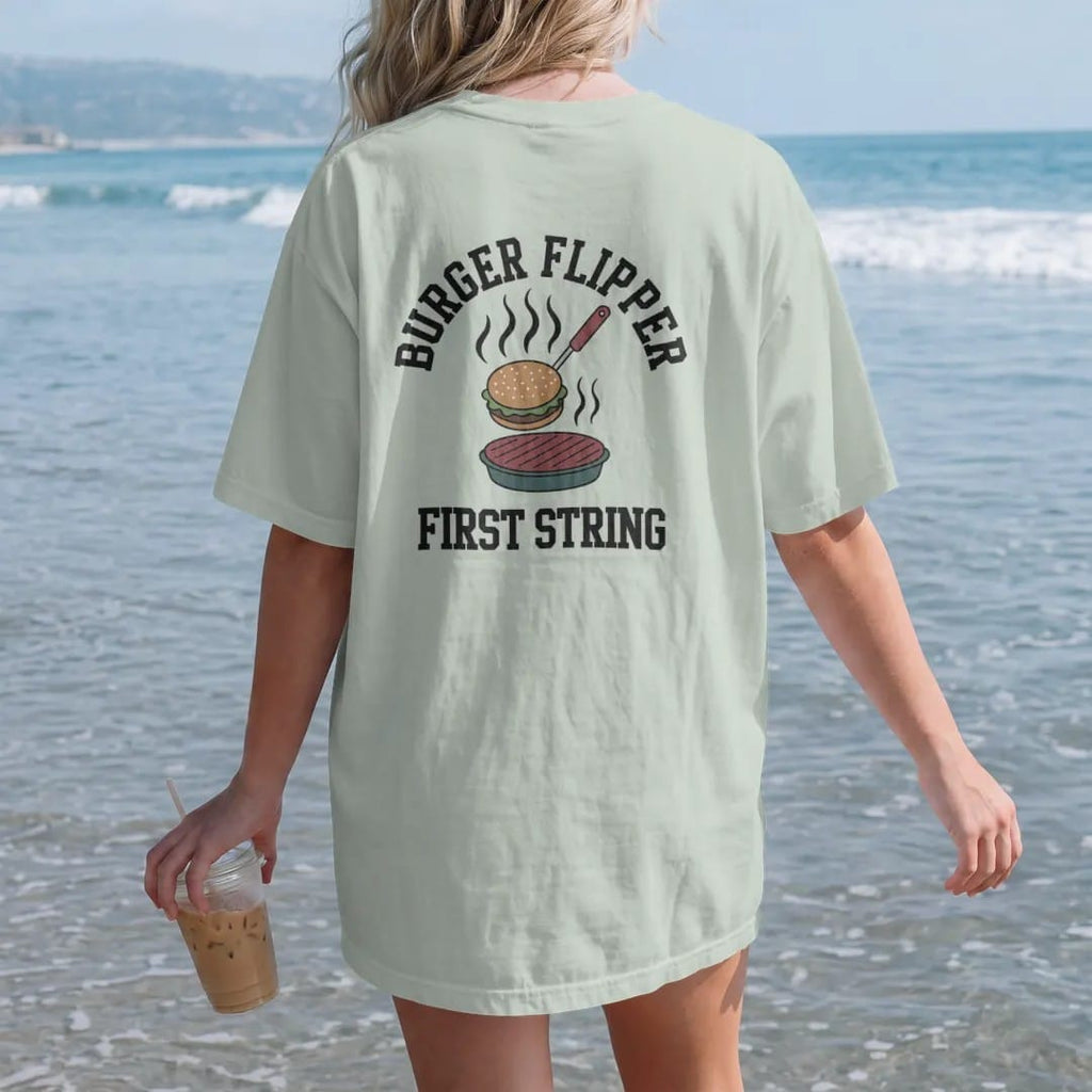Woman on the beach in a Bay tee, back view: a spatula flipping a burger patty into the air with grill smoke, with "BURGER FLIPPER" and "FIRST STRING" in chunky rounded 70s varsity letters, printed in faded stadium green, washed brick red, faded…