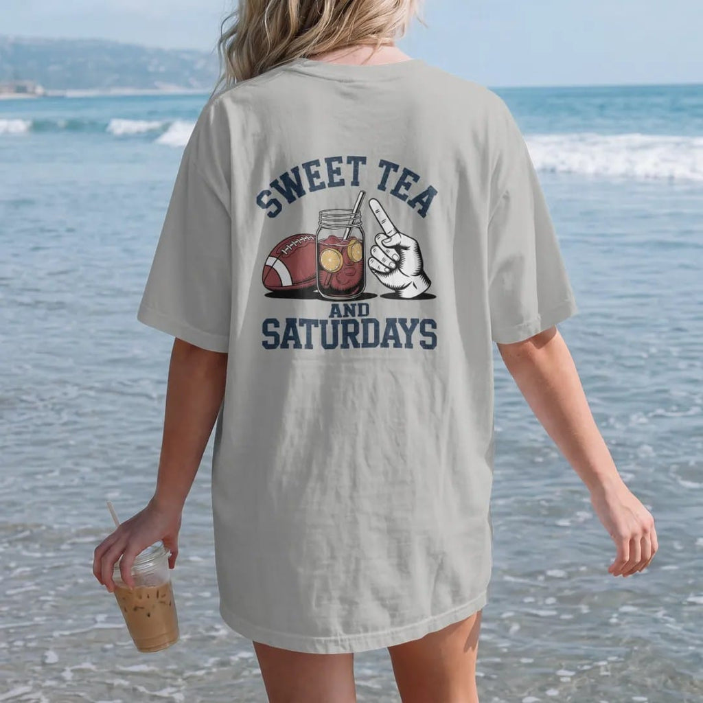 Woman on the beach in a Granite tee, back view: a mason jar of iced tea with lemon next to a football and a paper hand fan, with "SWEET TEA" and "AND SATURDAYS" in chunky 1970s swash script, printed in faded stadium green, washed brick red, faded…