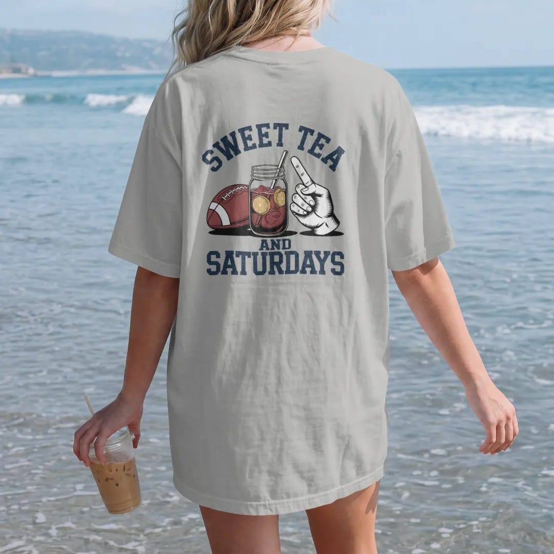 Woman on the beach in a Granite tee, back view: a mason jar of iced tea with lemon next to a football and a paper hand fan, with "SWEET TEA" and "AND SATURDAYS" in chunky 1970s swash script, printed in faded stadium green, washed brick red, faded…