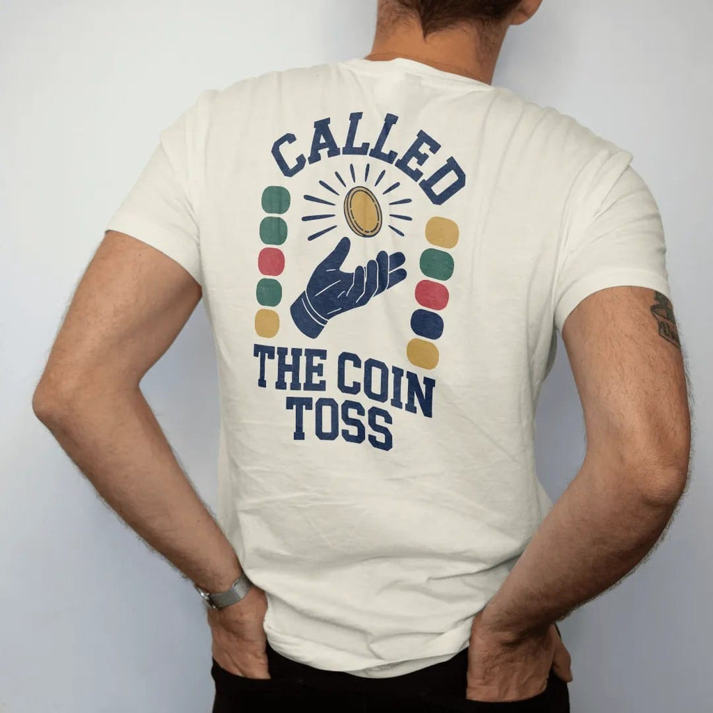 Man wearing a Ivory tee, seen from the back: a coin flipping in the air above a gloved hand with motion arcs, with "CALLED" and "THE COIN TOSS" in chunky 70s slab-serif letters, printed in faded stadium green, washed brick red, faded navy and…