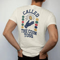 Man wearing a Ivory tee, seen from the back: a coin flipping in the air above a gloved hand with motion arcs, with "CALLED" and "THE COIN TOSS" in chunky 70s slab-serif letters, printed in faded stadium green, washed brick red, faded navy and…