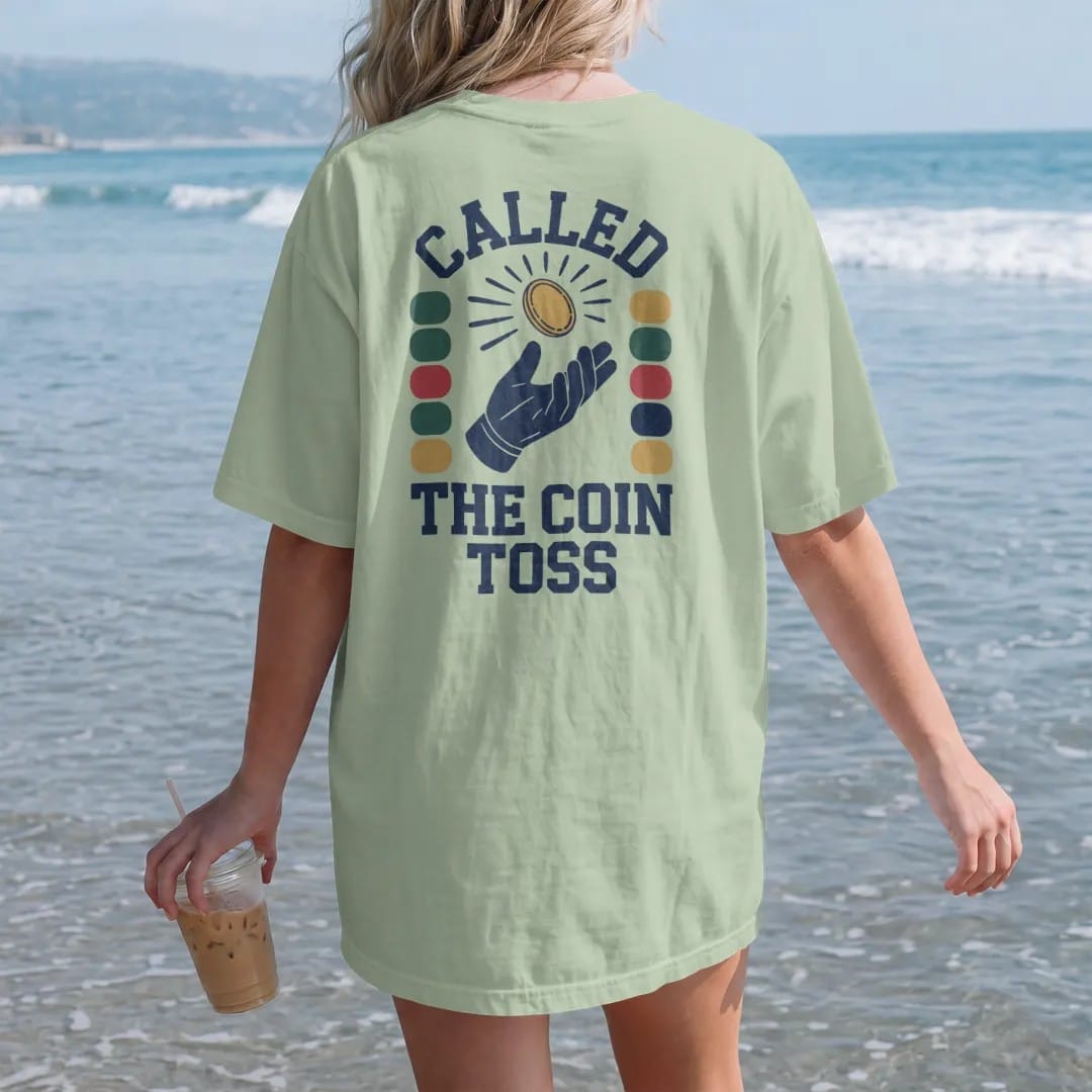 Woman on the beach in a Light Green tee, back view: a coin flipping in the air above a gloved hand with motion arcs, with "CALLED" and "THE COIN TOSS" in chunky 70s slab-serif letters, printed in faded stadium green, washed brick red, faded navy…