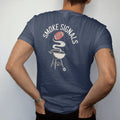 Man wearing a Navy tee, seen from the back: a kettle grill with smoke rising into the shape of a football, with "SMOKE SIGNALS" in chunky 1970s swash script, printed in faded bleacher cream, mustard, washed tomato red and sage field green with a…