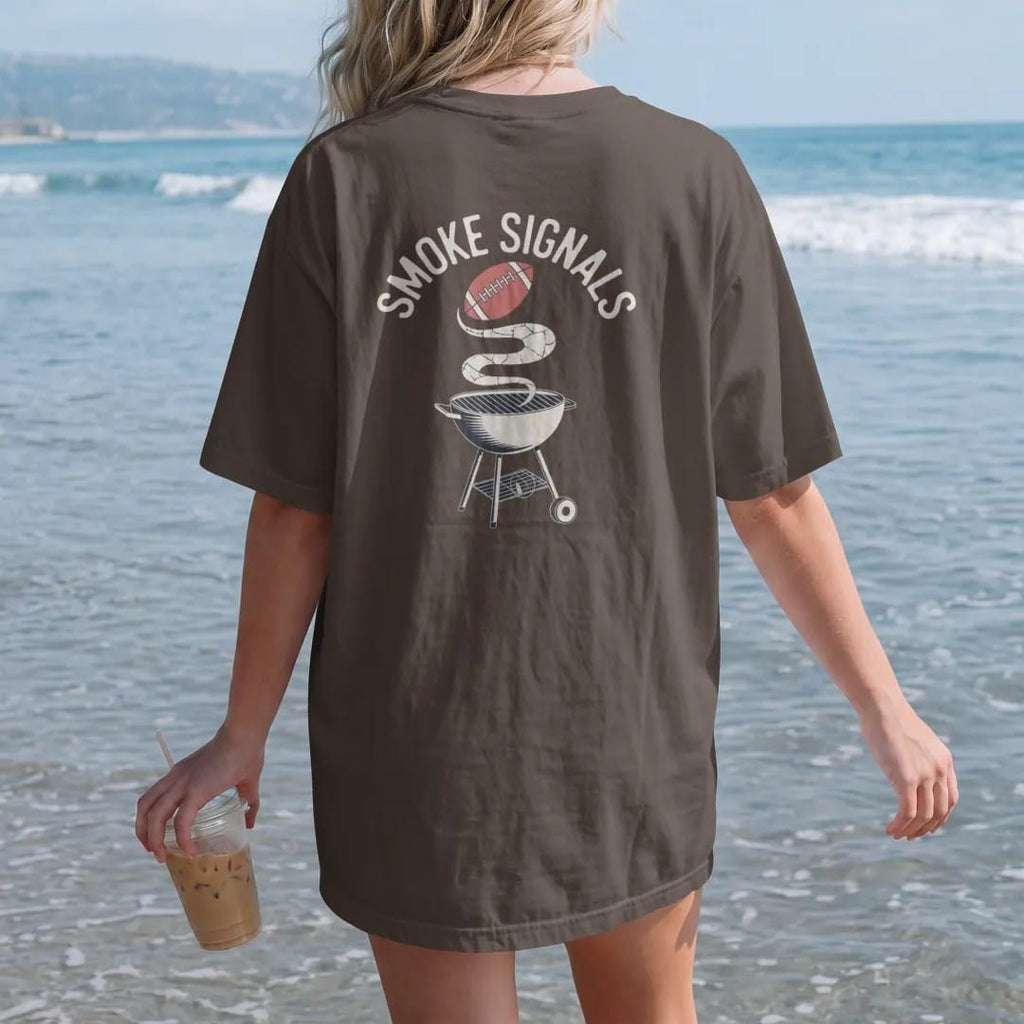 Woman on the beach in a Espresso tee, back view: a kettle grill with smoke rising into the shape of a football, with "SMOKE SIGNALS" in chunky 1970s swash script, printed in faded bleacher cream, mustard, washed tomato red and sage field green with…