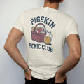Man wearing a Ivory tee, seen from the back: a gingham picnic basket with a football, sandwiches and a jug of lemonade, with "PIGSKIN" and "PICNIC CLUB" in chunky 1970s swash script, printed in faded stadium green, washed brick red, faded navy and…