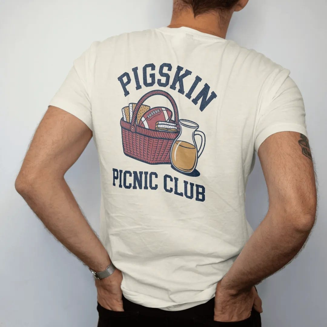 Man wearing a Ivory tee, seen from the back: a gingham picnic basket with a football, sandwiches and a jug of lemonade, with "PIGSKIN" and "PICNIC CLUB" in chunky 1970s swash script, printed in faded stadium green, washed brick red, faded navy and…