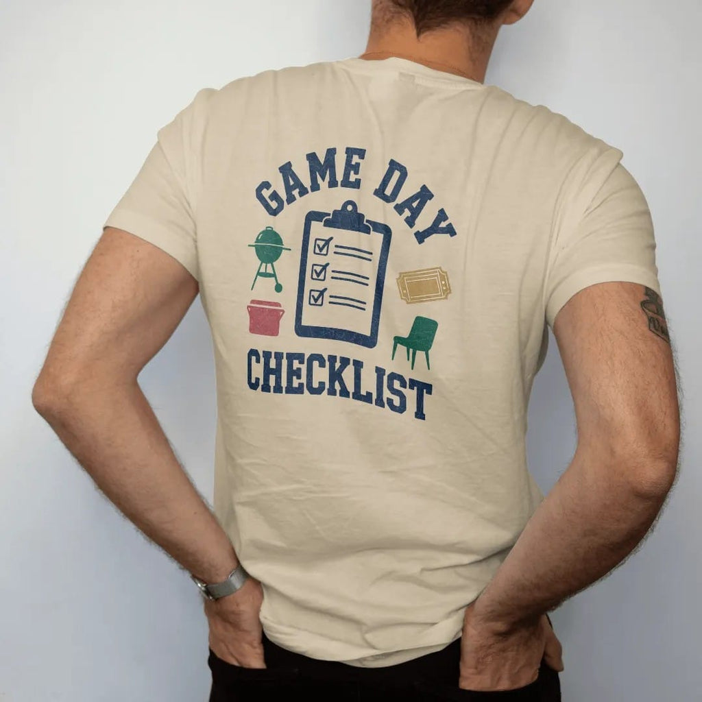 Man wearing a Sandstone tee, seen from the back: a clipboard with checked boxes next to icons of a grill, cooler, chair and ticket, with "GAME DAY" and "CHECKLIST" in chunky 70s slab-serif letters, printed in faded stadium green, washed brick red,…