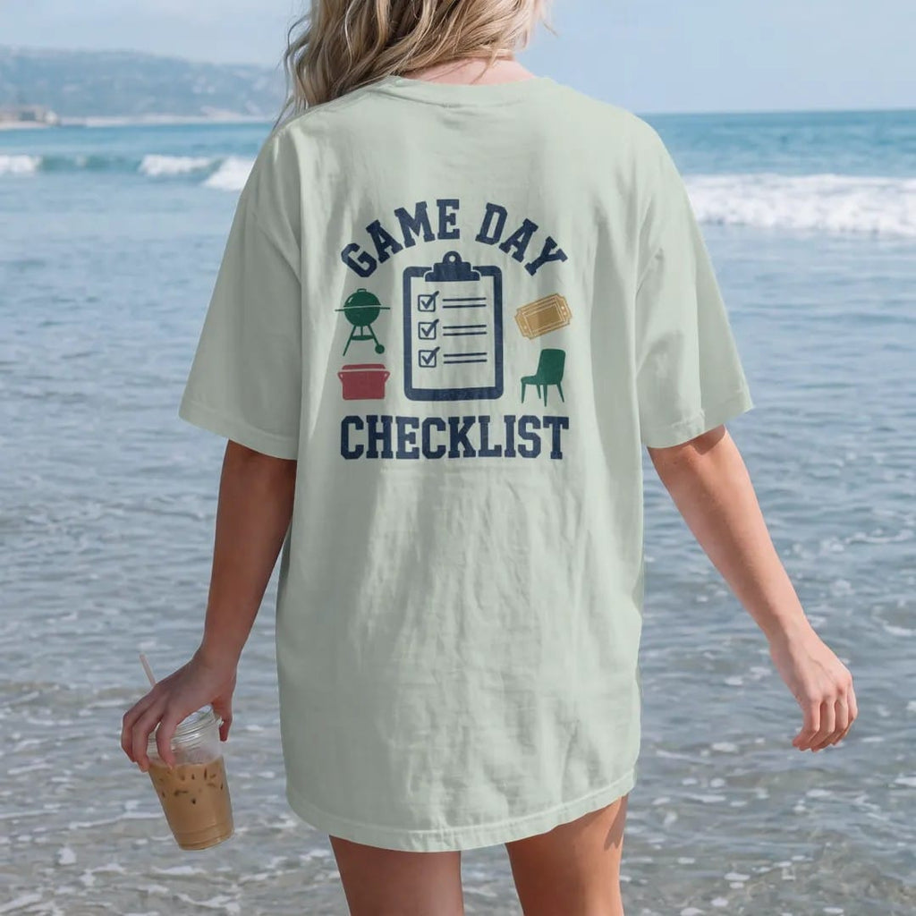 Woman on the beach in a Bay tee, back view: a clipboard with checked boxes next to icons of a grill, cooler, chair and ticket, with "GAME DAY" and "CHECKLIST" in chunky 70s slab-serif letters, printed in faded stadium green, washed brick red, faded…