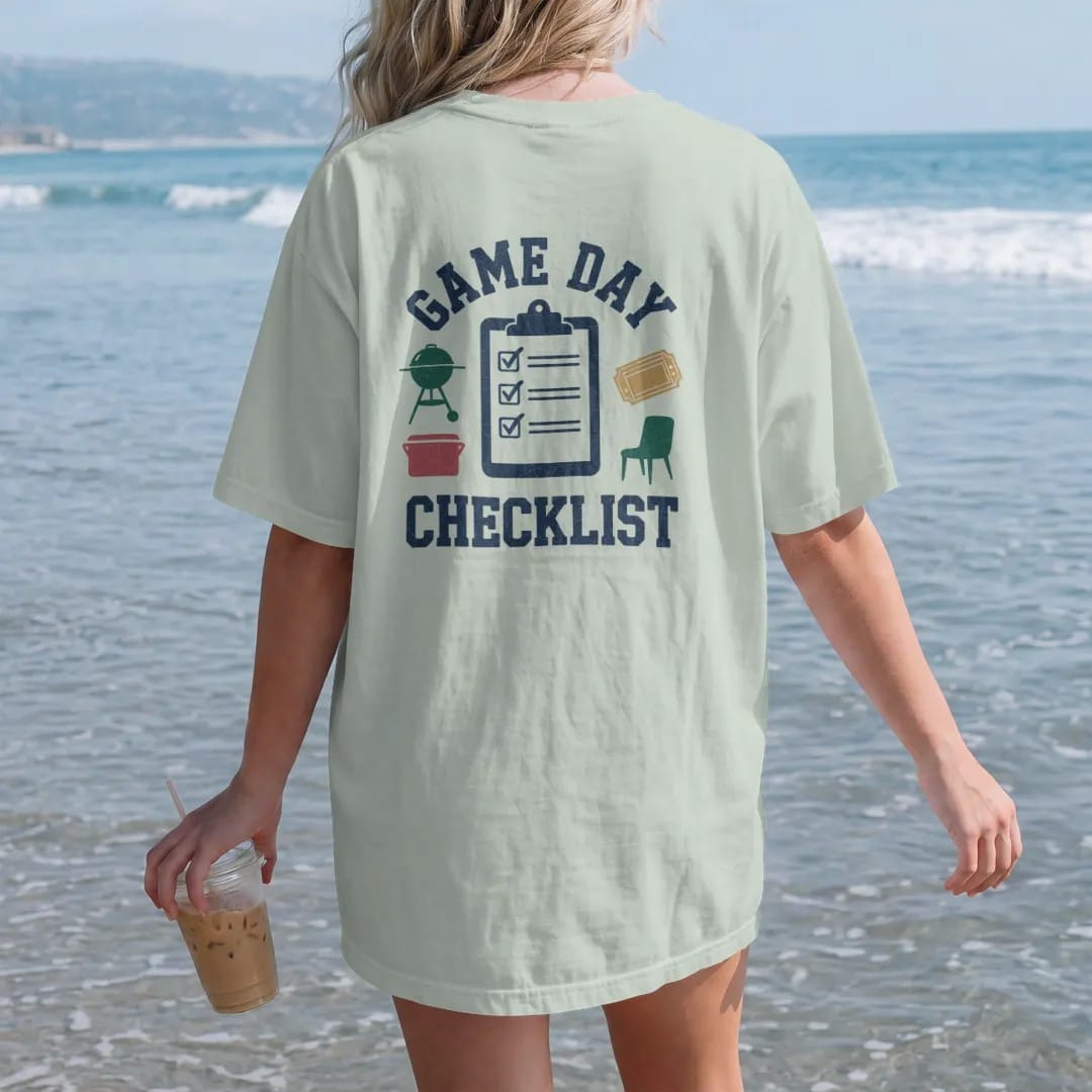 Woman on the beach in a Bay tee, back view: a clipboard with checked boxes next to icons of a grill, cooler, chair and ticket, with "GAME DAY" and "CHECKLIST" in chunky 70s slab-serif letters, printed in faded stadium green, washed brick red, faded…