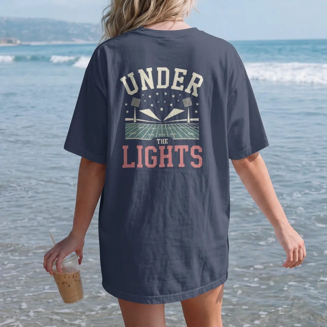 Woman on the beach in a Navy tee, back view: stadium floodlights blazing over a football field at night with stars, with "UNDER" and "THE LIGHTS" in chunky rounded 70s varsity letters, printed in faded bleacher cream, mustard, washed tomato red and…