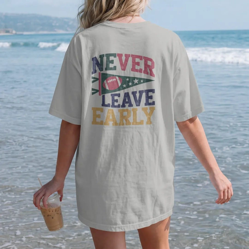 Woman on the beach in a Granite tee, back view: a felt pennant with a football and stars, with "NEVER LEAVE EARLY" in chunky rounded 70s varsity letters, printed in faded stadium green, washed brick red, faded navy and mustard with a cracked, worn…
