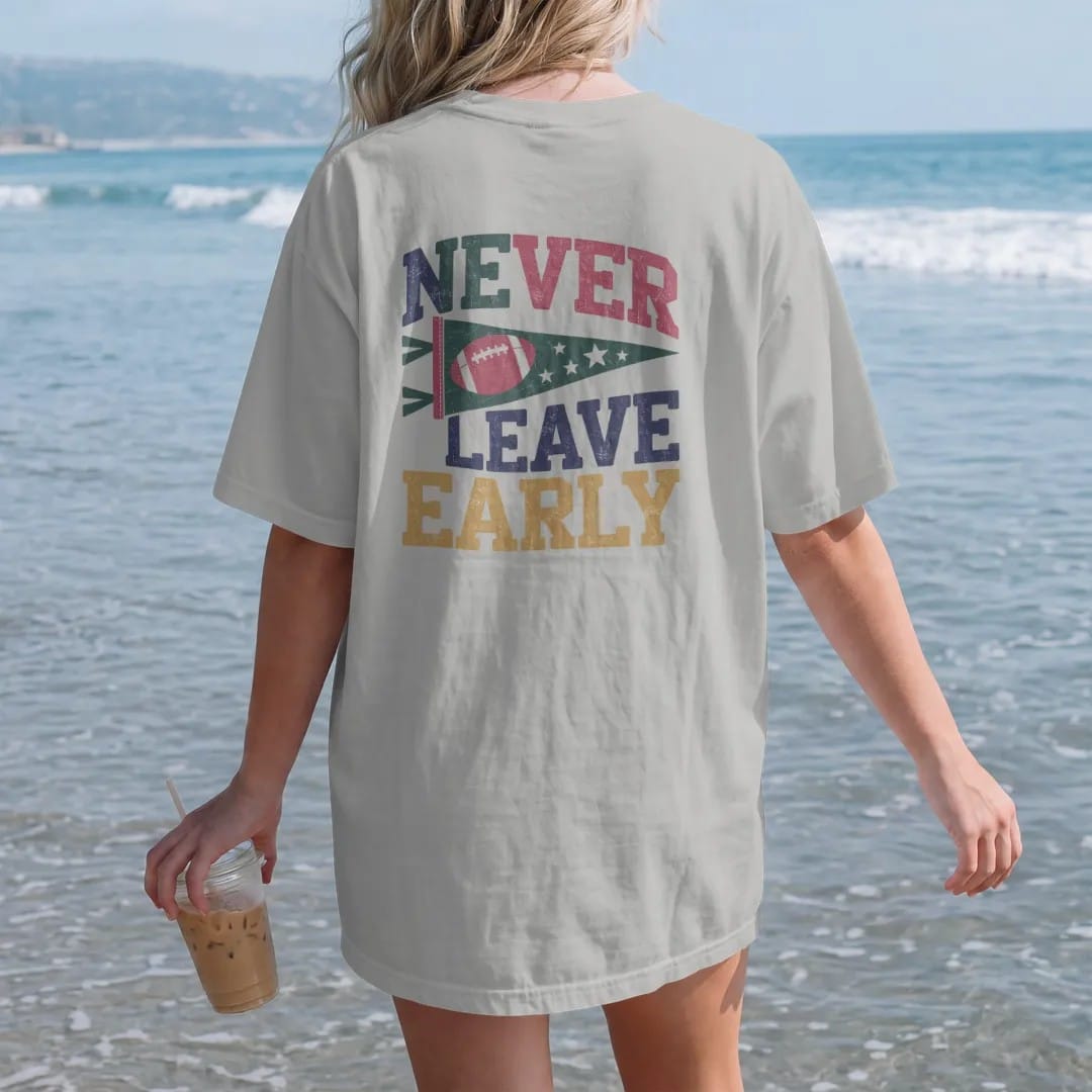 Woman on the beach in a Granite tee, back view: a felt pennant with a football and stars, with "NEVER LEAVE EARLY" in chunky rounded 70s varsity letters, printed in faded stadium green, washed brick red, faded navy and mustard with a cracked, worn…