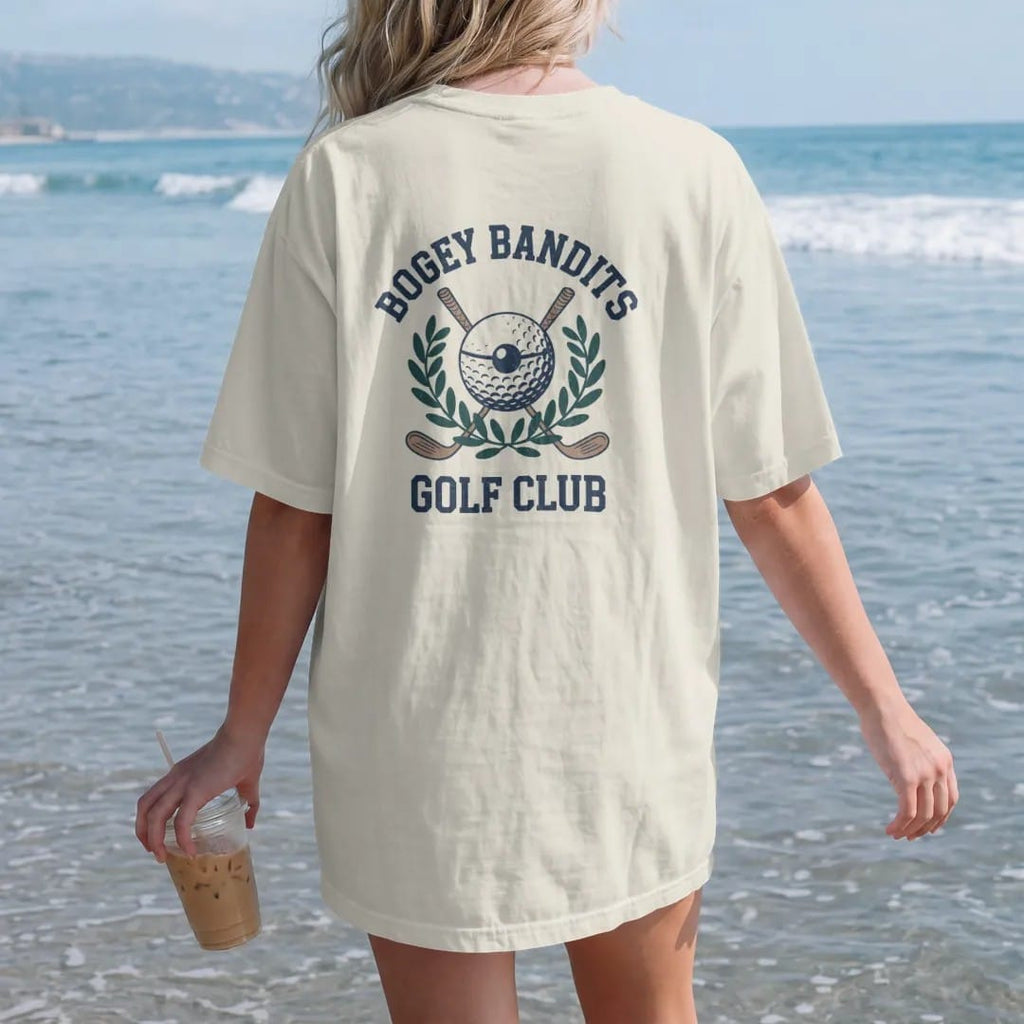 Woman on the beach in a Ivory tee, back view: a golf ball in a bandit eye mask over crossed clubs and a laurel wreath, with "BOGEY BANDITS" and "GOLF CLUB" in chunky rounded 70s serif letters, printed in faded fairway green, washed navy, faded flag…
