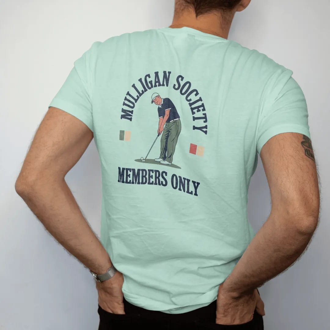 Man wearing a Chalky Mint tee, seen from the back: a golfer re-teeing a ball with a sly grin, with "MULLIGAN SOCIETY" and "MEMBERS ONLY" in chunky rounded 70s serif letters, printed in faded fairway green, washed navy, faded flag red and sand tan…