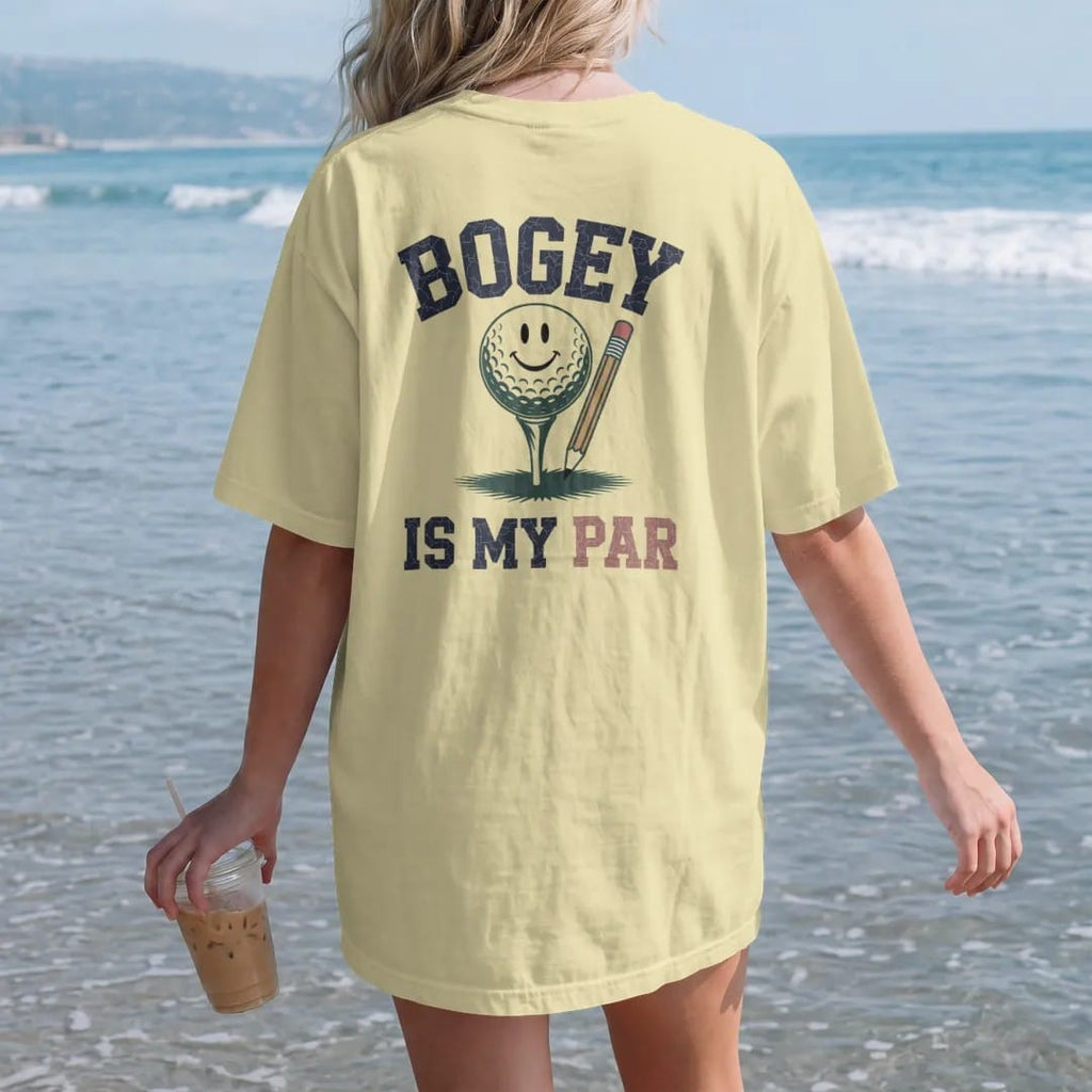 Woman on the beach in a Butter tee, back view: a ball resting on a tee next to a stubby scorecard pencil, with a smiley face on the ball, with "BOGEY" and "IS MY PAR" in chunky faded collegiate block letters, printed in faded fairway green, washed…