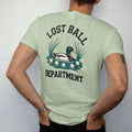 Man wearing a Light Green tee, seen from the back: a duck paddling in a pond surrounded by floating golf balls and reeds, with "LOST BALL" and "DEPARTMENT" in chunky rounded 70s serif letters, printed in faded fairway green, washed navy, faded flag…