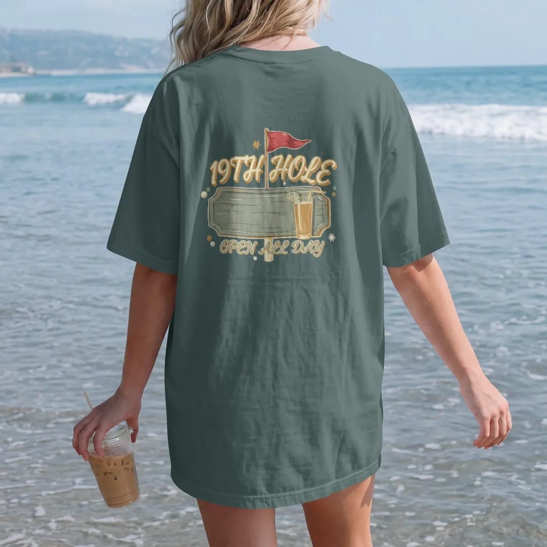 Woman on the beach in a Blue Spruce tee, back view: a vintage clubhouse bar sign with a flagstick and a frosty drink, with "19TH HOLE" and "OPEN ALL DAY" in loopy 1970s script, printed in faded clubhouse cream, sage grass green, washed flag red and…