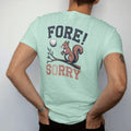 Man wearing a Chalky Mint tee, seen from the back: a golf ball flying toward a startled squirrel on a branch, with "FORE!" and "SORRY" in chunky faded collegiate block letters, printed in faded fairway green, washed navy, faded flag red and sand…