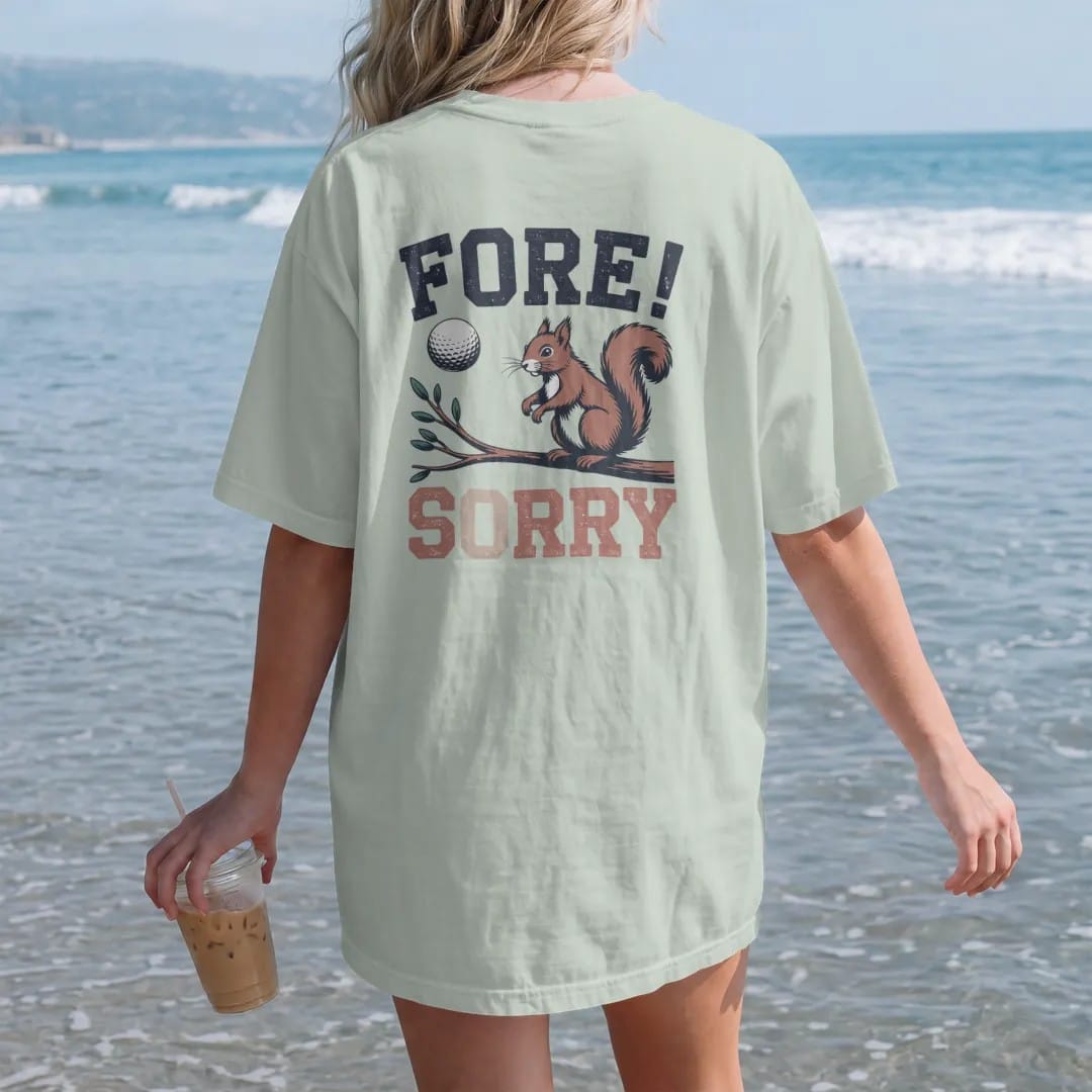 Woman on the beach in a Bay tee, back view: a golf ball flying toward a startled squirrel on a branch, with "FORE!" and "SORRY" in chunky faded collegiate block letters, printed in faded fairway green, washed navy, faded flag red and sand tan with…