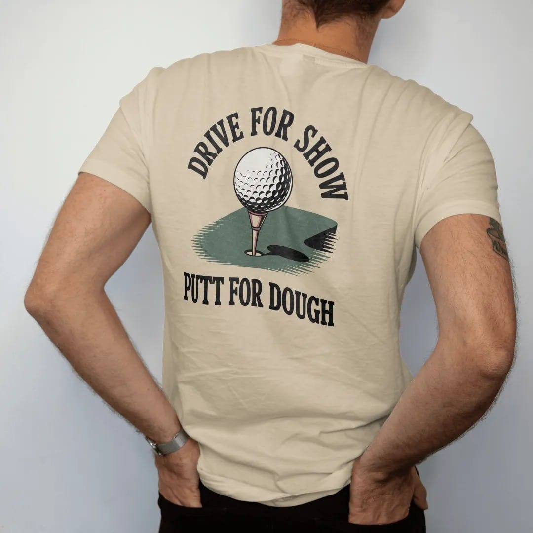 Man wearing a Sandstone tee, seen from the back: a golf ball rolling toward the cup on a sloping green, with "DRIVE FOR SHOW" and "PUTT FOR DOUGH" in loopy 1970s script, printed in faded fairway green, washed navy, faded flag red and sand tan with…
