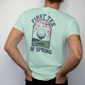 Man wearing a Chalky Mint tee, seen from the back: a ball on a tee with blooming cherry blossoms framing a green fairway, with "FIRST TEE" and "OF SPRING" in loopy 1970s script, printed in faded fairway green, washed navy, faded flag red and sand…