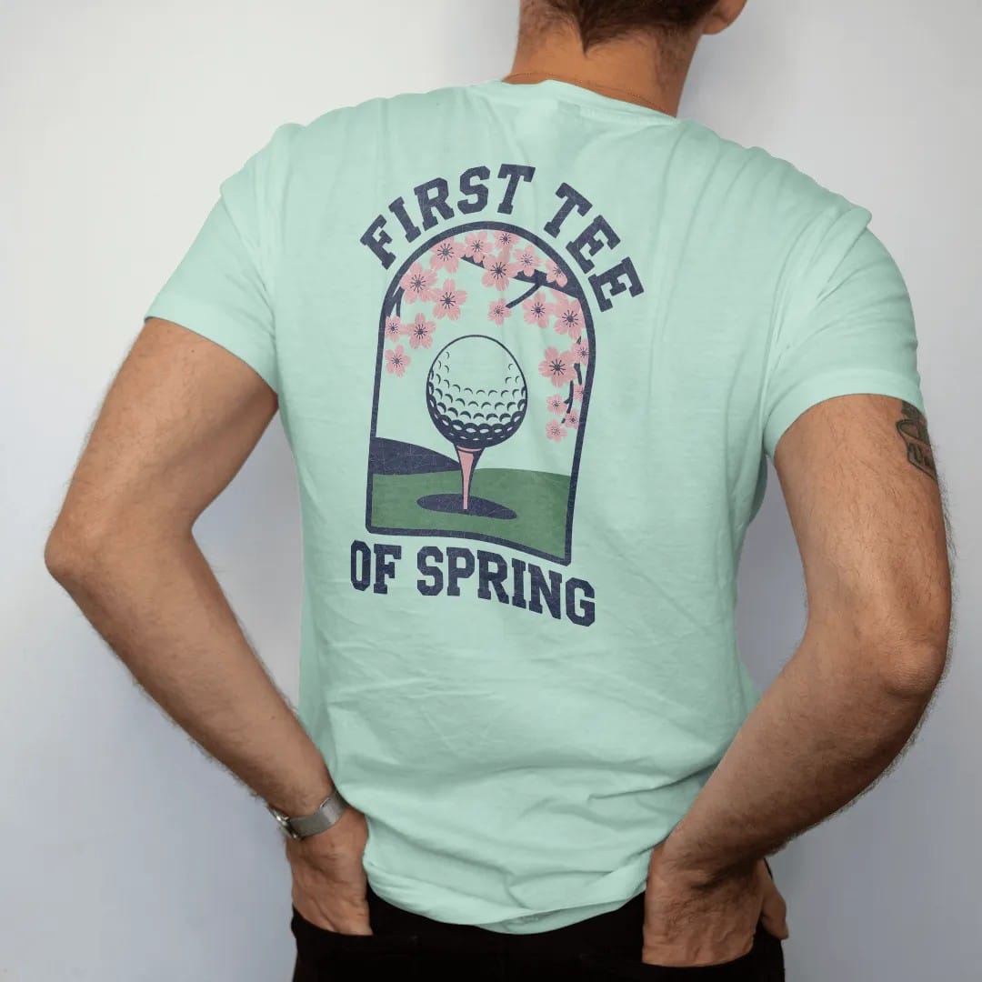 Man wearing a Chalky Mint tee, seen from the back: a ball on a tee with blooming cherry blossoms framing a green fairway, with "FIRST TEE" and "OF SPRING" in loopy 1970s script, printed in faded fairway green, washed navy, faded flag red and sand…
