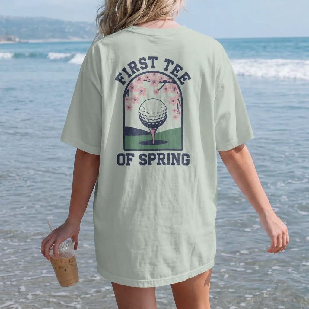 Woman on the beach in a Bay tee, back view: a ball on a tee with blooming cherry blossoms framing a green fairway, with "FIRST TEE" and "OF SPRING" in loopy 1970s script, printed in faded fairway green, washed navy, faded flag red and sand tan with…