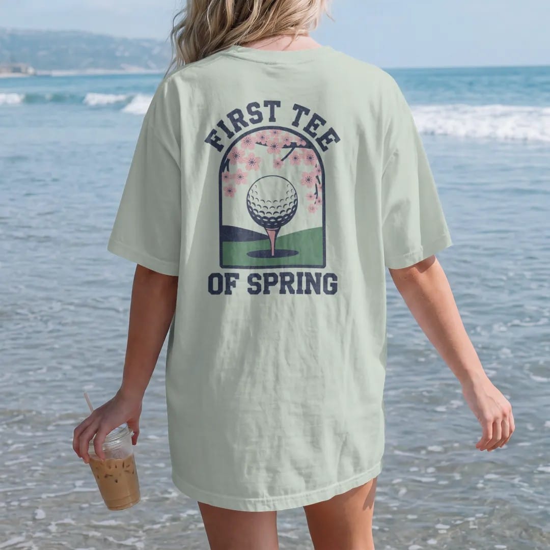 Woman on the beach in a Bay tee, back view: a ball on a tee with blooming cherry blossoms framing a green fairway, with "FIRST TEE" and "OF SPRING" in loopy 1970s script, printed in faded fairway green, washed navy, faded flag red and sand tan with…