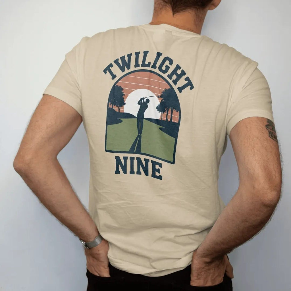 Man wearing a Sandstone tee, seen from the back: a golfer silhouetted on a fairway at sunset with long shadows, with "TWILIGHT" and "NINE" in chunky rounded 70s serif letters, printed in faded fairway green, washed navy, faded flag red and sand tan…