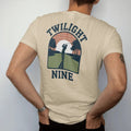 Man wearing a Sandstone tee, seen from the back: a golfer silhouetted on a fairway at sunset with long shadows, with "TWILIGHT" and "NINE" in chunky rounded 70s serif letters, printed in faded fairway green, washed navy, faded flag red and sand tan…