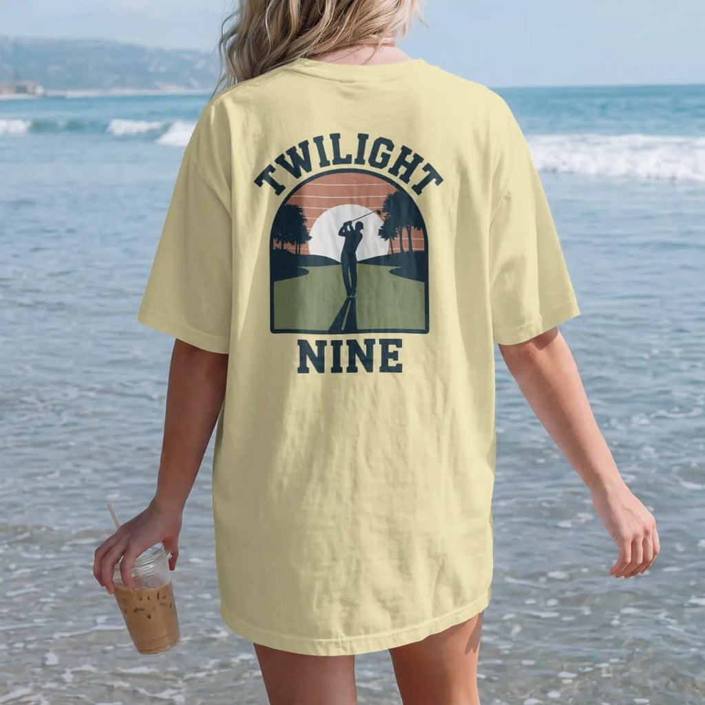 Woman on the beach in a Butter tee, back view: a golfer silhouetted on a fairway at sunset with long shadows, with "TWILIGHT" and "NINE" in chunky rounded 70s serif letters, printed in faded fairway green, washed navy, faded flag red and sand tan…