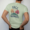 Man wearing a Light Green tee, seen from the back: a fairway lined with orange and red trees, leaves drifting onto the green, with "FALL" and "FAIRWAYS" in loopy 1970s script, printed in faded fairway green, washed navy, faded flag red and sand tan…