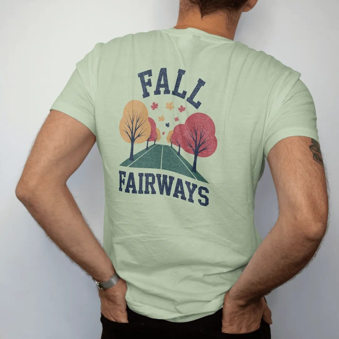 Man wearing a Light Green tee, seen from the back: a fairway lined with orange and red trees, leaves drifting onto the green, with "FALL" and "FAIRWAYS" in loopy 1970s script, printed in faded fairway green, washed navy, faded flag red and sand tan…