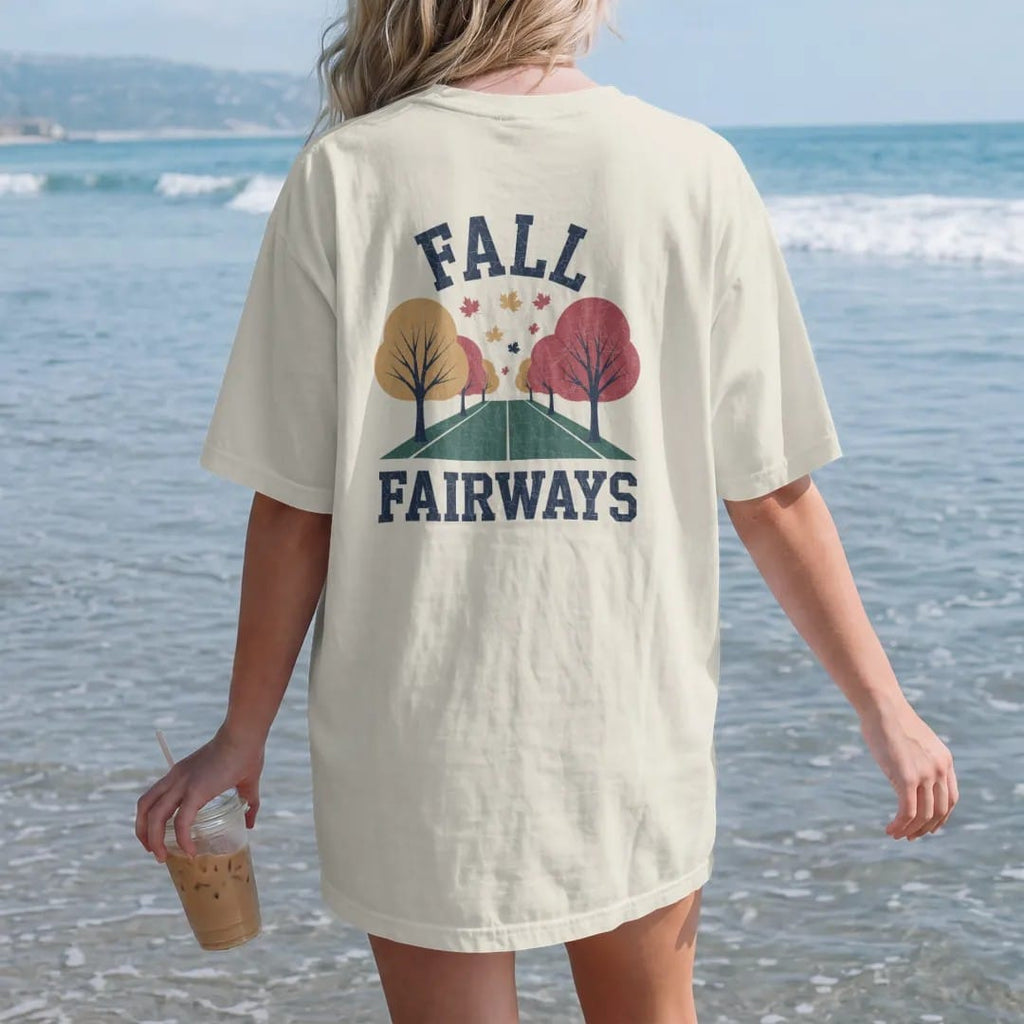 Woman on the beach in a Ivory tee, back view: a fairway lined with orange and red trees, leaves drifting onto the green, with "FALL" and "FAIRWAYS" in loopy 1970s script, printed in faded fairway green, washed navy, faded flag red and sand tan with…
