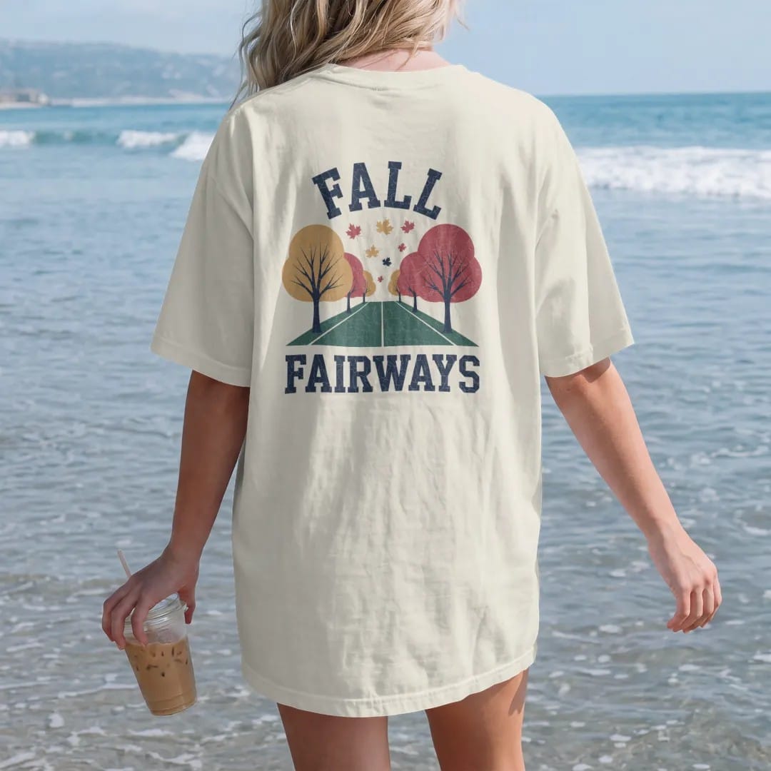 Woman on the beach in a Ivory tee, back view: a fairway lined with orange and red trees, leaves drifting onto the green, with "FALL" and "FAIRWAYS" in loopy 1970s script, printed in faded fairway green, washed navy, faded flag red and sand tan with…