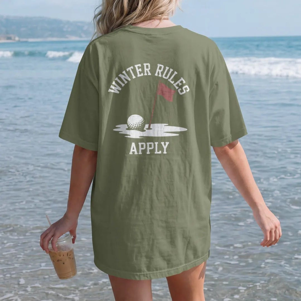 Woman on the beach in a Moss tee, back view: a golf ball half-buried in snow next to a red flagstick, with "WINTER RULES" and "APPLY" in chunky faded collegiate block letters, printed in faded clubhouse cream, sage grass green, washed flag red and…