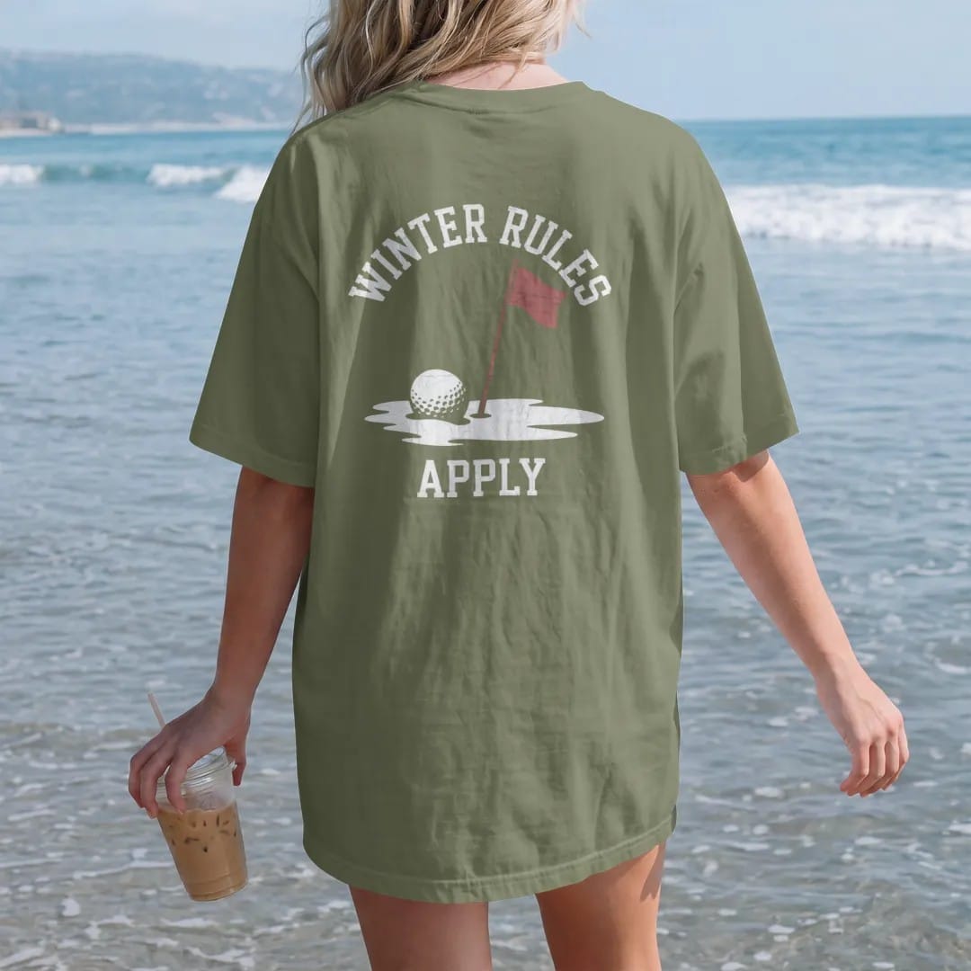 Woman on the beach in a Moss tee, back view: a golf ball half-buried in snow next to a red flagstick, with "WINTER RULES" and "APPLY" in chunky faded collegiate block letters, printed in faded clubhouse cream, sage grass green, washed flag red and…